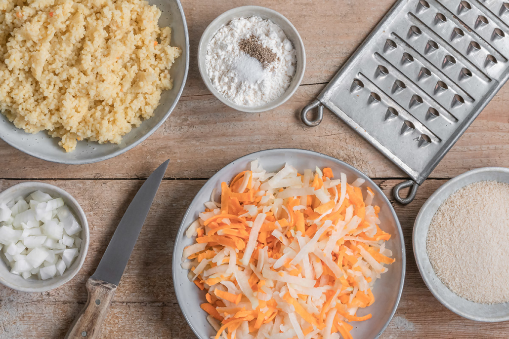 Hirse, Mehl, Salz, Pfeffer, Zwiebeln, Paniermehl sowie Raspeln aus Kohlrabi und Karotten in verschiedenen Schüsseln daneben liegt eine Reibe und ein Messer