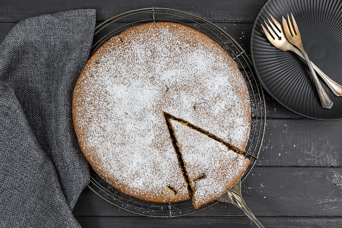 Angeschnittener Rührkuchen mit Äpfeln und Zimt auf einem Kuchengitter. Ein Stück wird mit dem Kuchenheber herausgezogen. Ein Teller mit zwei Kuchengabeln steht bereit.